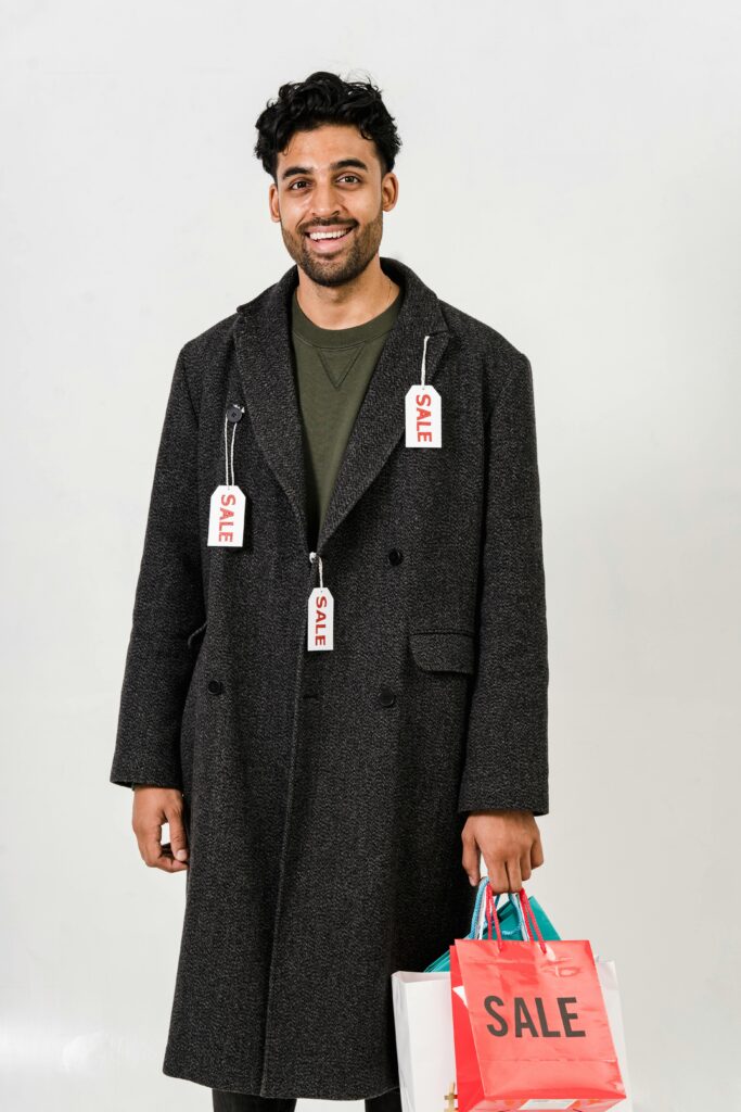 Smiling man wearing an overcoat with sale tags, holding a shopping bag during a promotional event.