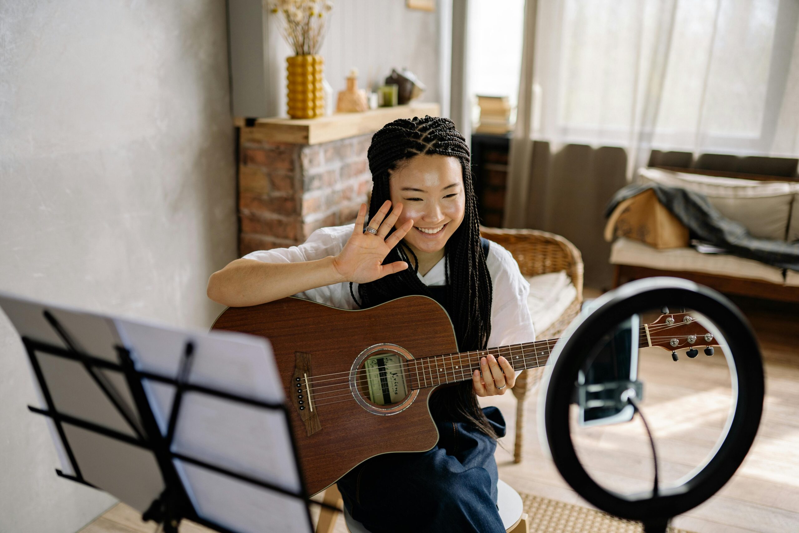 Smiling woman streaming acoustic guitar session from home using smartphone and ring light.