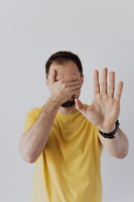Man in yellow shirt covering face with one hand while showing a stop gesture with the other.