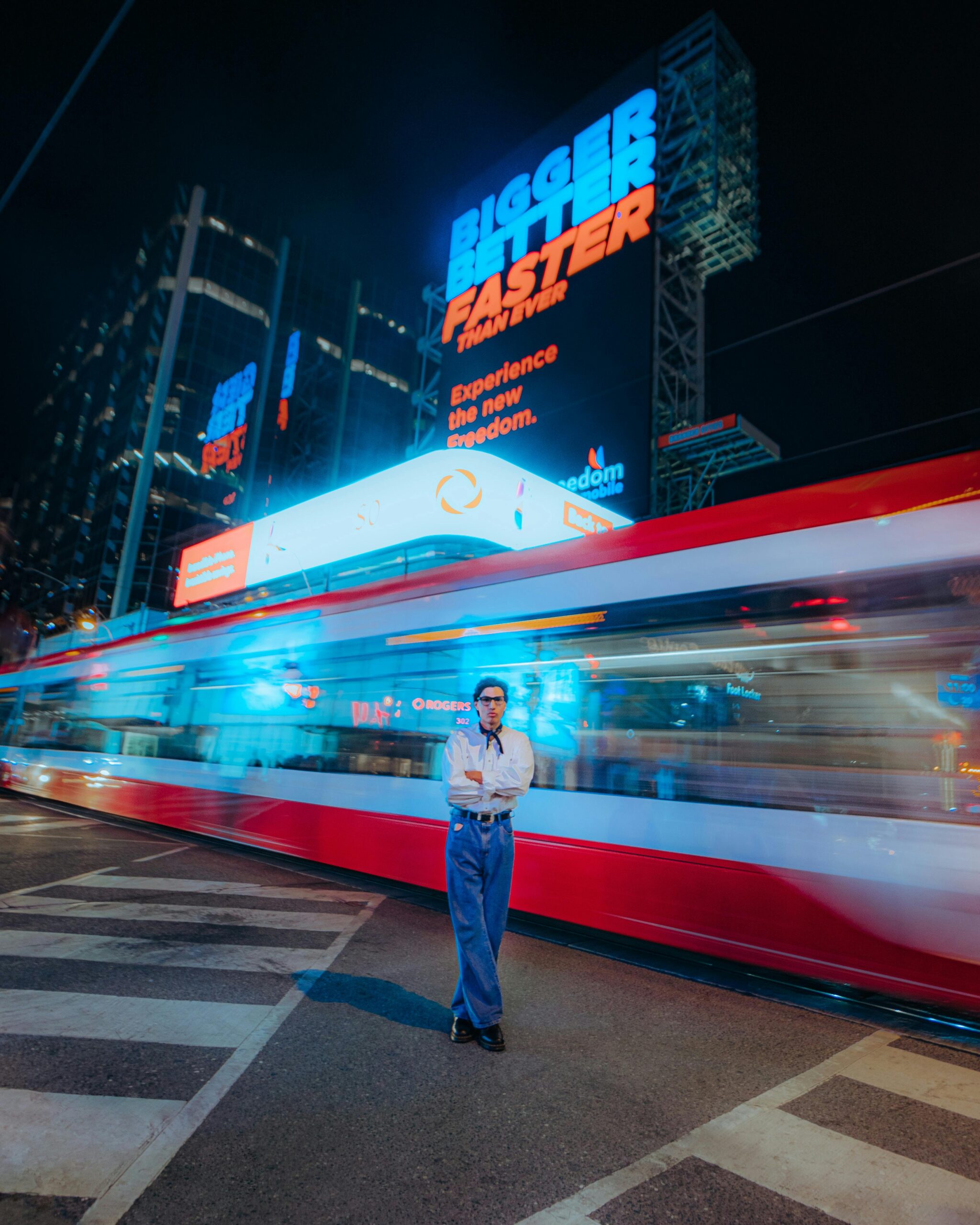 A person standing confidently on city streets at night with a passing tram in motion.